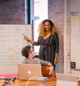 a smiling woman stands in front of a white board as a colleague turns in her chair to talk about how to hire a freelance writer